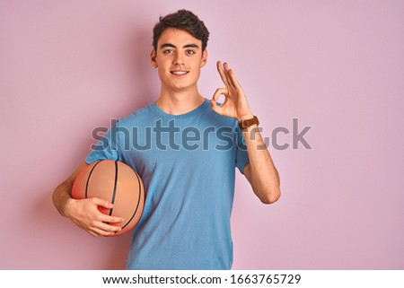 Teenager boy holding professional basket ball over isolated pink background doing ok sign with fingers, excellent symbol