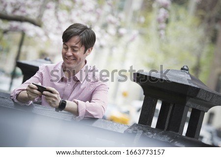 City life in spring. A man sitting outdoors in a city park. Looking down at his smart phone and smiling.