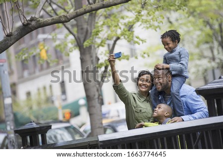 A new york city, park in the spring. A boy riding on his father's shoulders, and a woman using a smart phone taking a selfy picture.