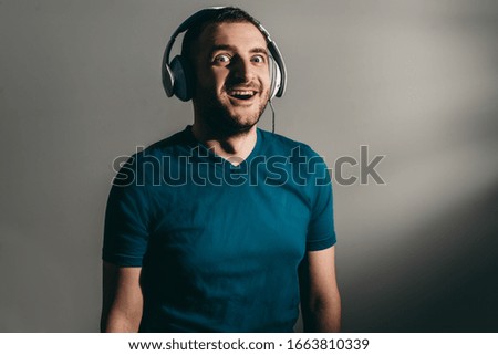 Young bearded crazy emotion man with white headphones listens to music on dark gray background in nightclub.