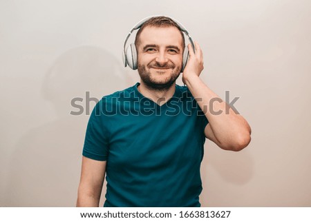 Young bearded emotion smiles man with white headphones listens to music on light background .