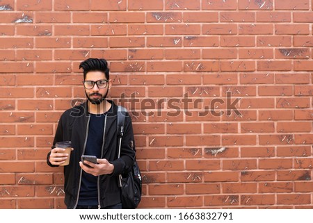 Young indian student man with phone and coffee cup standing against brick wall