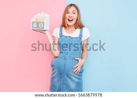 The 40 weeks of pregnancy. A stunned young beautiful pregnant woman poses holding a sign with the term of pregnancy, standing on an isolated pink and blue background waiting for the birth of a child.