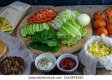 Preparing for the preparation of hamburgers. On a wooden board are lettuce leaves, chopped: tomatoes, cucumbers, onions, cutlets and sauces.