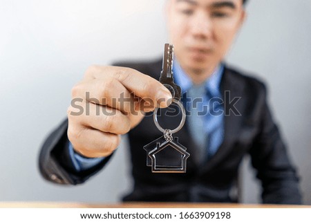 concept of a male asian sales businessman holding a key with a key ring of a house representing a real-estate agent, selling house, home insurance, rent, and wearing a suit with grey flat background  