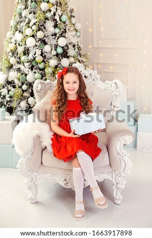 Happy funny child girl in a red dress on the background of a Christmas tree with a gift in his hands. Little girl picture in the New Year decorations. New Year's advertising. 