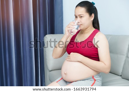 Young Asian pregnant woman holding  glass of water. Healthy living. Her sitting in liveing room. Nutrition and diet during pregnancy.