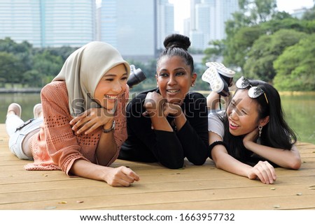 Three woman Malay Chinese Indian Asian Malaysian outdoor green park lake nature lying on front wooden deck hand on cheek talk smile chat emotion expression Royalty-Free Stock Photo #1663957732