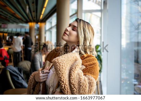 Beautiful European woman wearing brown fall fashion clothing is happily relaxing and enjoying a look outside the window in the city. the female model has brown blonde highlights and siting indoor