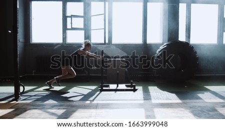 Sliding side view young athletic Caucasian woman exercising with heavy training sled in big atmospheric gym slow motion.