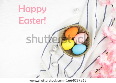Pastel colored eggs in the nest with the branch of pink flowers on white wooden background. Easter holiday. Text Happy Easter in the corner of picture.