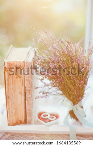 Platinum or silver wedding rings on the heart shaped white plate with pink heather flowers, vintage book and wedding bouquet at sunset lights