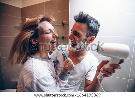 Beautiful young couple in love having fun playing with hair dryer in the bathroom. Husband and wife laughing together at home.