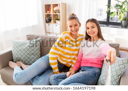 people, technology and friendship concept - happy teenage girls taking picture with selfie stick sitting on sofa at home