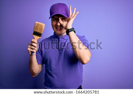 Young modern decorator painter man holding paint brush over purple background with happy face smiling doing ok sign with hand on eye looking through fingers