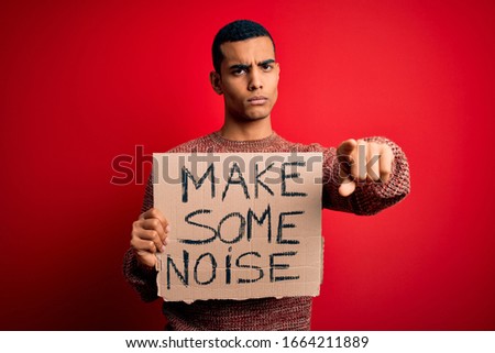Handsome african american activist man protesting holding banner with make noise message pointing with finger to the camera and to you, hand sign, positive and confident gesture from the front