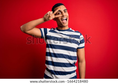 Handsome african american man wearing casual striped t-shirt standing over red background Doing peace symbol with fingers over face, smiling cheerful showing victory