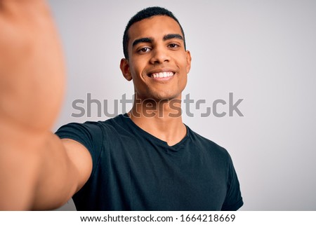 Young handsome african american man wearing casual t-shirt making selfie by camera with a happy face standing and smiling with a confident smile showing teeth