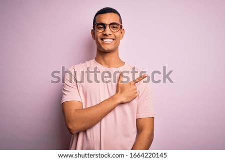 Handsome african american man wearing casual t-shirt and glasses over pink background cheerful with a smile on face pointing with hand and finger up to the side with happy and natural expression