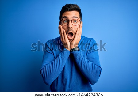 Young handsome man with beard wearing casual sweater and glasses over blue background afraid and shocked, surprise and amazed expression with hands on face Royalty-Free Stock Photo #1664224366