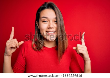 Young beautiful brunette girl wearing casual t-shirt over isolated red background smiling amazed and surprised and pointing up with fingers and raised arms.