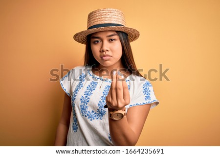 Young beautiful asian girl wearing casual t-shirt and hat standing over yellow background Doing Italian gesture with hand and fingers confident expression