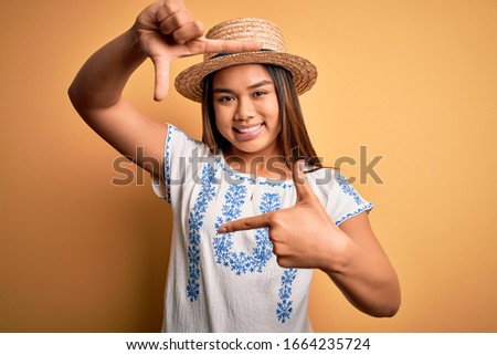 Young beautiful asian girl wearing casual t-shirt and hat standing over yellow background smiling making frame with hands and fingers with happy face. Creativity and photography concept.