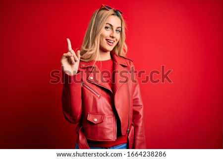 Young beautiful blonde woman wearing casual jacket standing over isolated red background showing and pointing up with finger number one while smiling confident and happy.