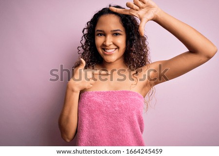 Young beautiful woman with curly hair wearing shower towel after bath over pink background smiling making frame with hands and fingers with happy face. Creativity and photography concept.
