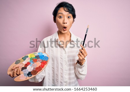 Young beautiful chinese artist woman painting using paintbrush and palette with colors scared in shock with a surprise face, afraid and excited with fear expression