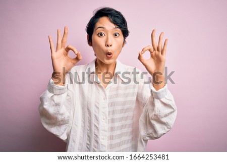 Young beautiful asian girl wearing casual shirt standing over isolated pink background looking surprised and shocked doing ok approval symbol with fingers. Crazy expression