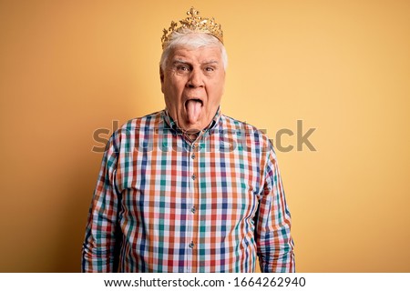 Senior handsome hoary man wearing golden crown of king over isolated yellow background sticking tongue out happy with funny expression. Emotion concept.