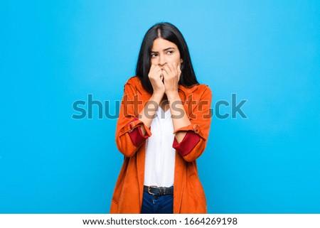 young pretty latin woman looking worried, anxious, stressed and afraid, biting fingernails and looking to lateral copy space against flat wall