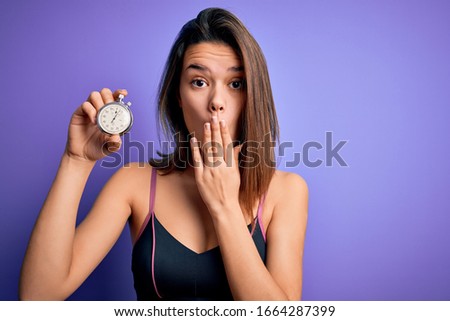 Young beautiful sporty girl doing sport using stopwatch over isolated purple background cover mouth with hand shocked with shame for mistake, expression of fear, scared in silence, secret concept