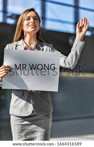Portrait of businesswoman waving for her Chinese boss while holding blank white board in arrival area at airport