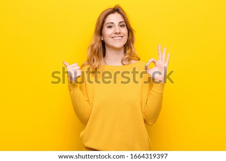 young pretty blonde woman feeling happy, amazed, satisfied and surprised, showing okay and thumbs up gestures, smiling against yellow wall