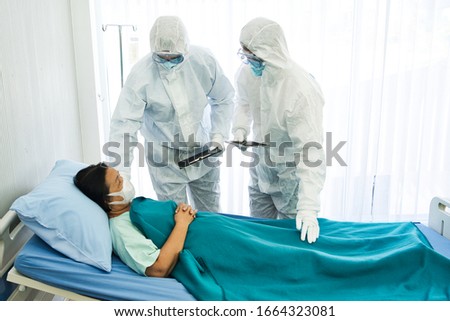 Two doctors in the personal protective suits examining the Coronavirus COVID 19 infected aging female patient in the control area.  Royalty-Free Stock Photo #1664323081