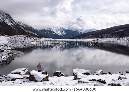 Staring into a Glassy Canadian Lake.

A lone man looks towards the snow capped mountains that tower above a picture perfect icy body of water.

Snow and rocks are in the foreground.