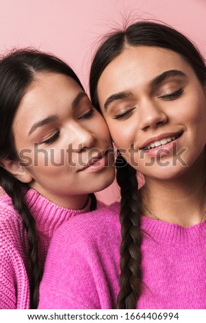 Image of two happy teenage girls with braids in casual clothes smiling at camera isolated over pink background