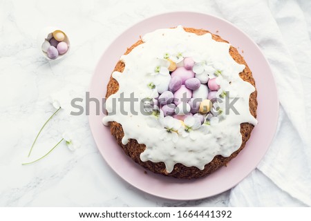 Glazed easter cake decorated with flowers and mini chocolate eggs candy on white marble background. Happy Easter Holidays. Top view. Copy space.