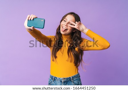 Image of young beautiful woman with long brown hair smiling and taking selfie photo on cellphone isolated over violet background