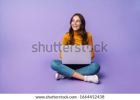 Image of young beautiful woman using laptop while sitting with legs crossed isolated over violet background
