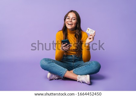 Image of young beautiful woman holding smartphone and credit card while sitting with legs crossed isolated over violet background