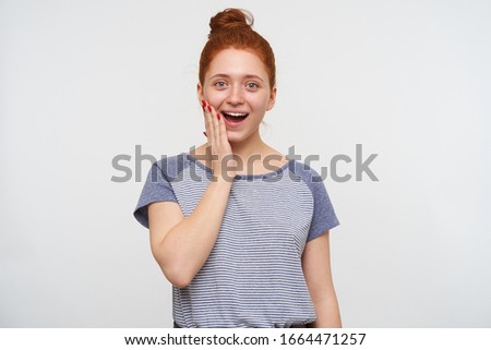 Surprised young beautiful redhead female with bun hairstyle holding raised palm on her cheek and looking excitedly at camera, standing against white background