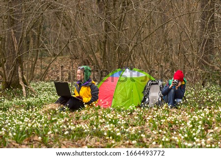 A tent in a forest with snowdrops, a family in spring resting on a country walk among wild trees and a large number of beautiful white fragrant flowers. Mom works on a laptop , old retro camera