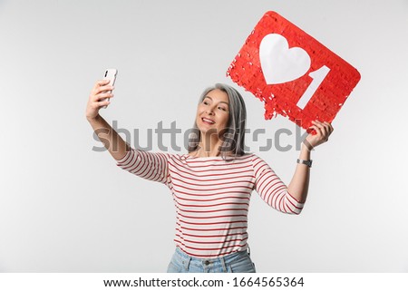 Image of adult mature woman with long white hair holding cellphone and heart like symbol on placard isolated over gray background