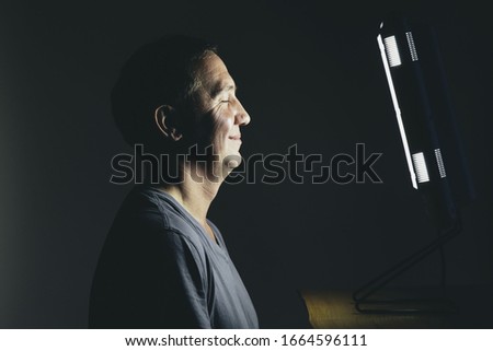 Smiling middle aged man sitting in front of a light therapy box, a full spectrum light box which mimics the sun, and treats people suffering from seasonal affective disorder.