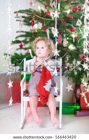 Beautiful little girl in a red dress checking her Christmas stocking sitting in a white rocking chair under a decorated tree