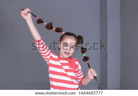 portrait of a young funny girl of seven years old with a funny hairstyle with a striped sweater in a good mood and grimaces on a gray background