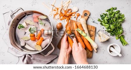 Cooking - chef's hands preparing chicken stock (broth or bouillon) with vegetables in a pot. Kitchen - grey concrete worktop scenery from above (top view, flat lay).